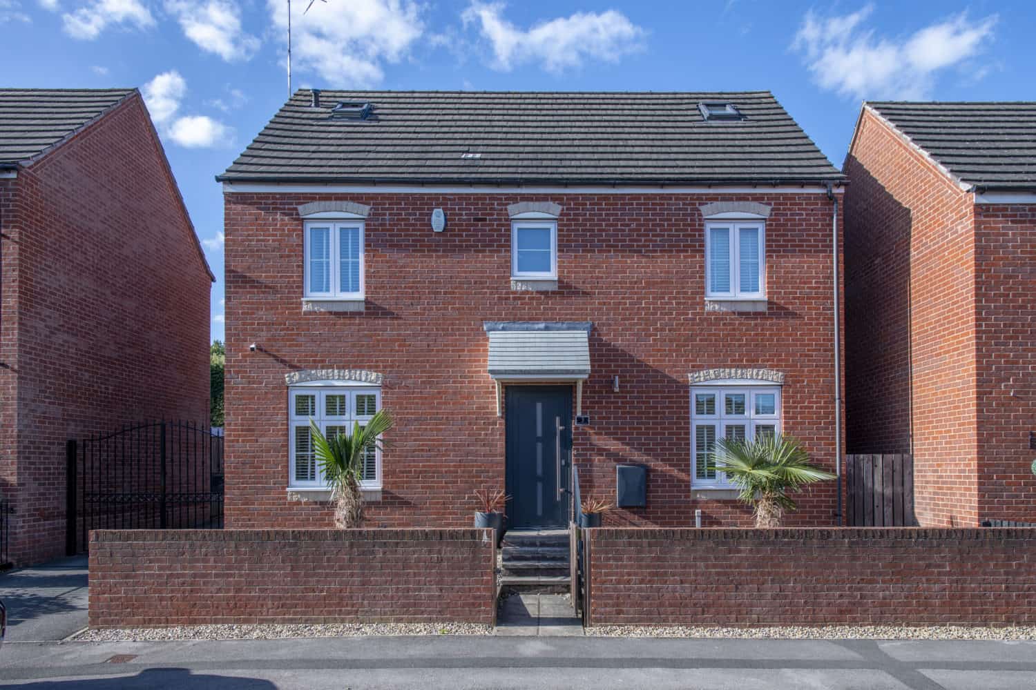 A modern two-story red-brick house with white-trimmed windows, a sleek dark front door, small palm trees in front, and a low brick wall bordering the property. The blue sky above features scattered clouds.