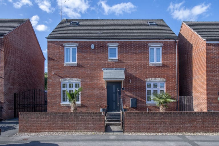 A modern two-story red-brick house with white-trimmed windows, a sleek dark front door, small palm trees in front, and a low brick wall bordering the property. The blue sky above features scattered clouds.