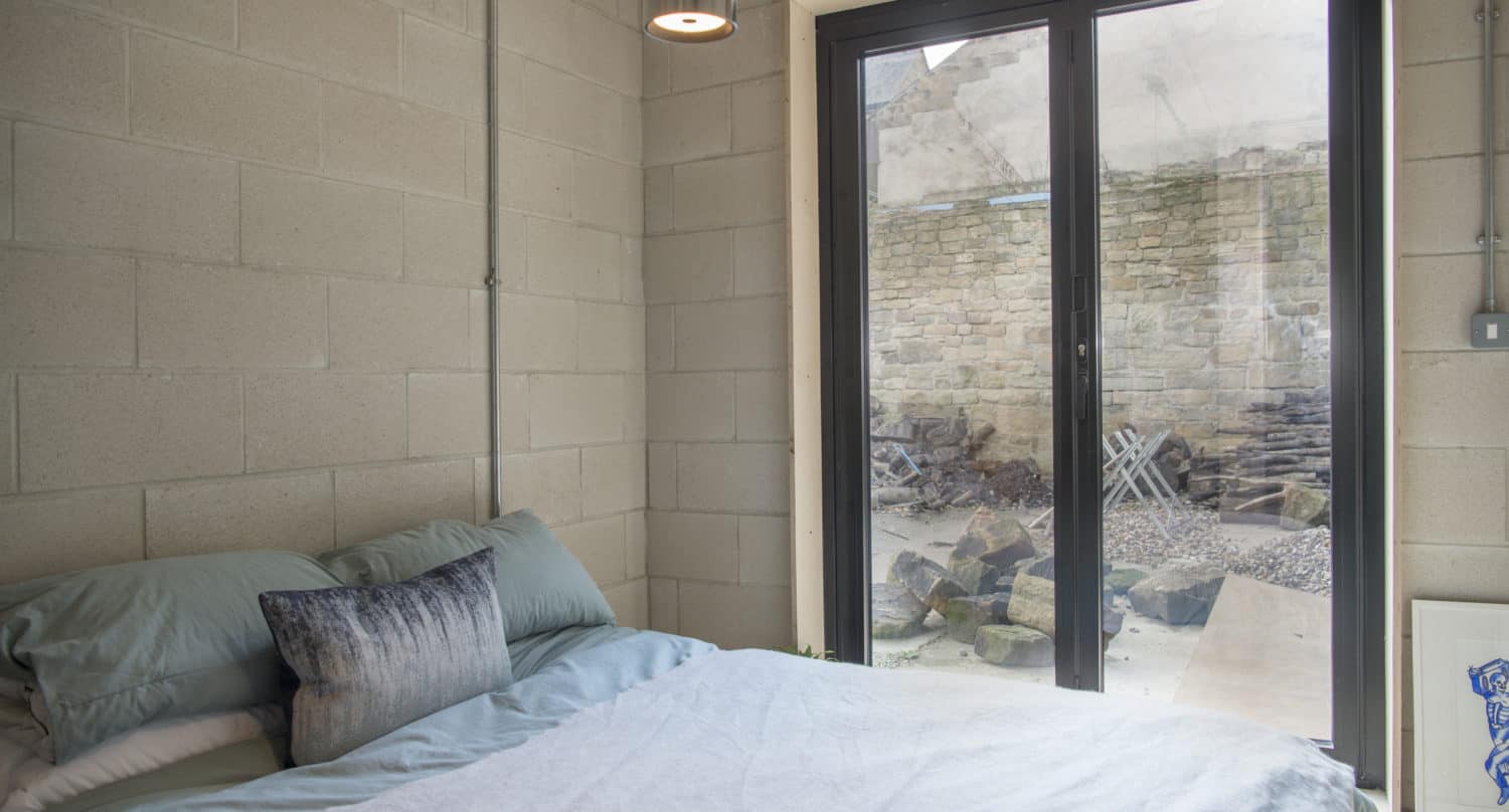 A modern bedroom with light-colored concrete block walls, a neatly made bed with blue and white bedding, a small pendant lamp, and large sliding doors opening to a stone patio with outdoor chairs and rocks.
