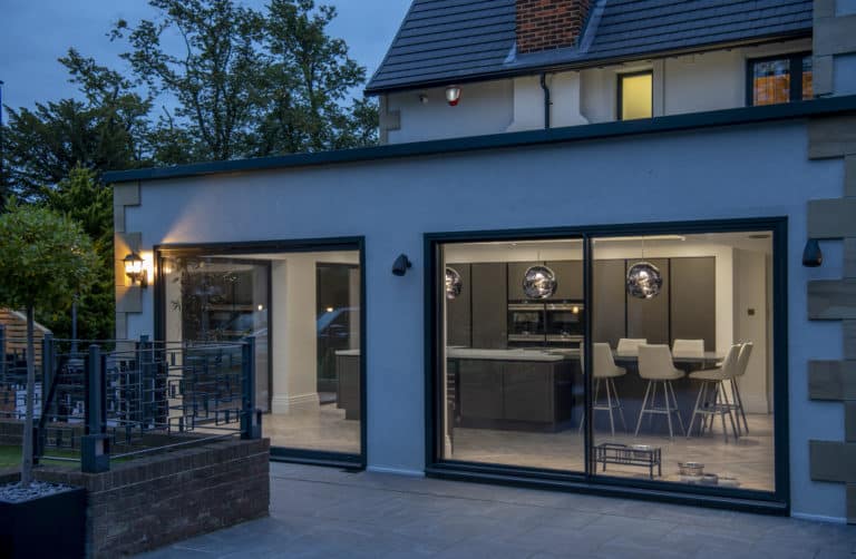 Modern house exterior at dusk featuring expansive bifold doors that reveal a contemporary kitchen and dining area with bar stools; outdoor lights warmly illuminate the inviting patio space.