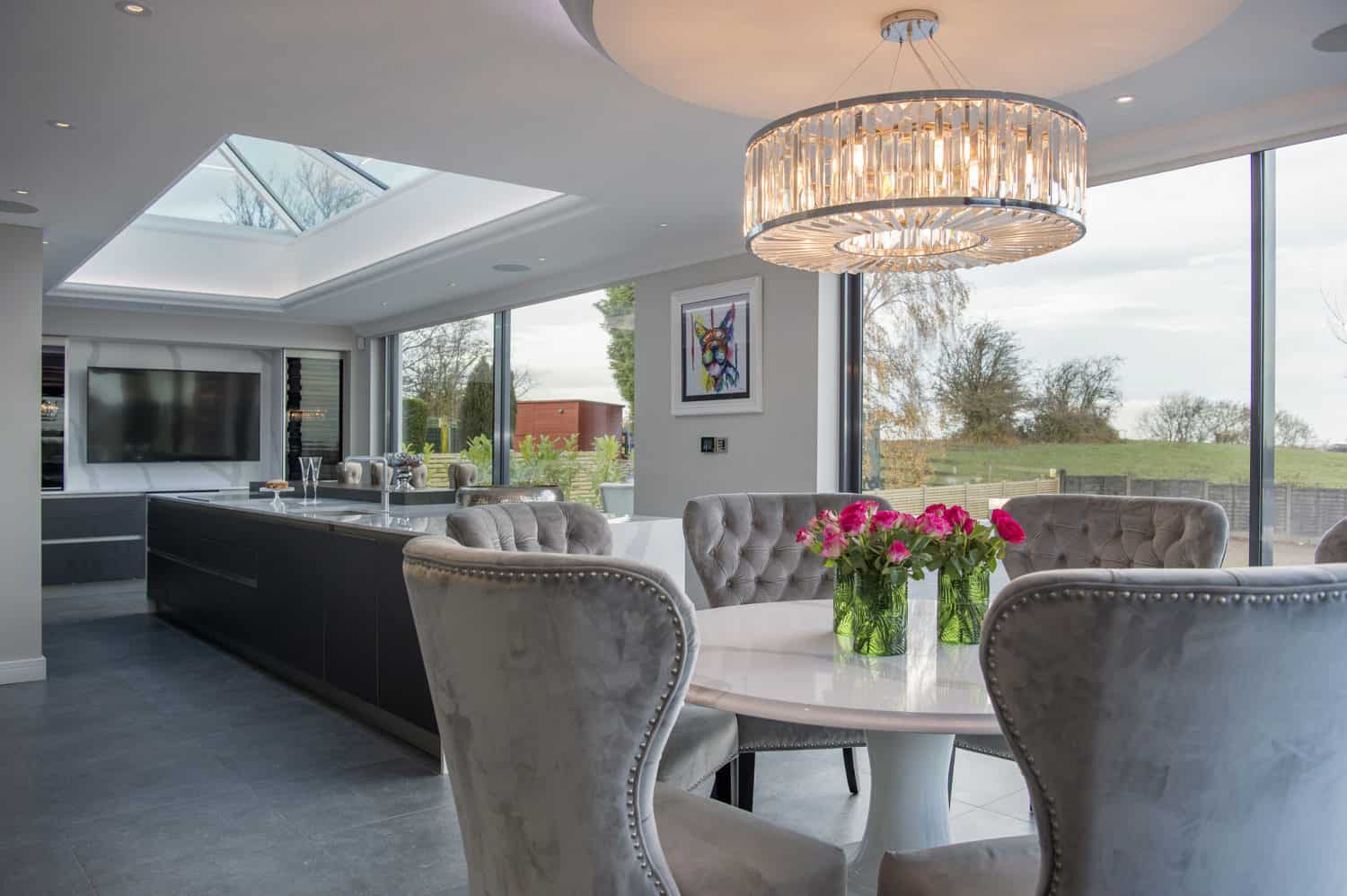 Modern open-plan kitchen and dining area with large windows, glass roofing, a round table surrounded by gray upholstered chairs, and a vase of pink flowers. A chandelier hangs above the table, and trees are visible outside.