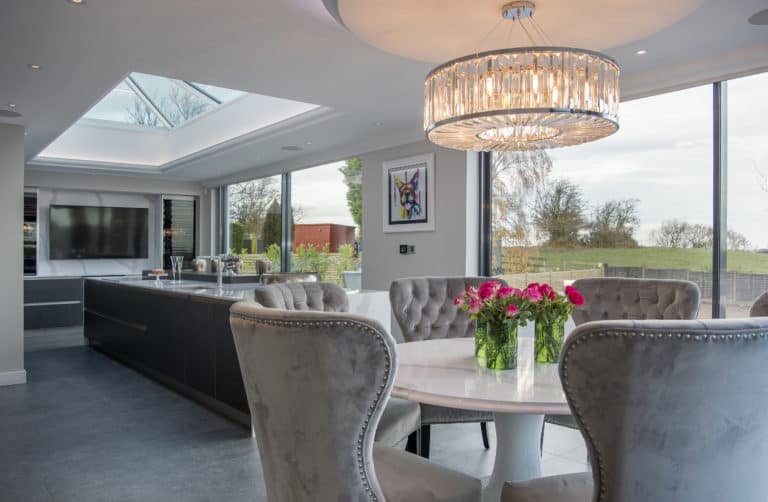 Modern open-plan kitchen and dining area with large windows, glass roofing, a round table surrounded by gray upholstered chairs, and a vase of pink flowers. A chandelier hangs above the table, and trees are visible outside.