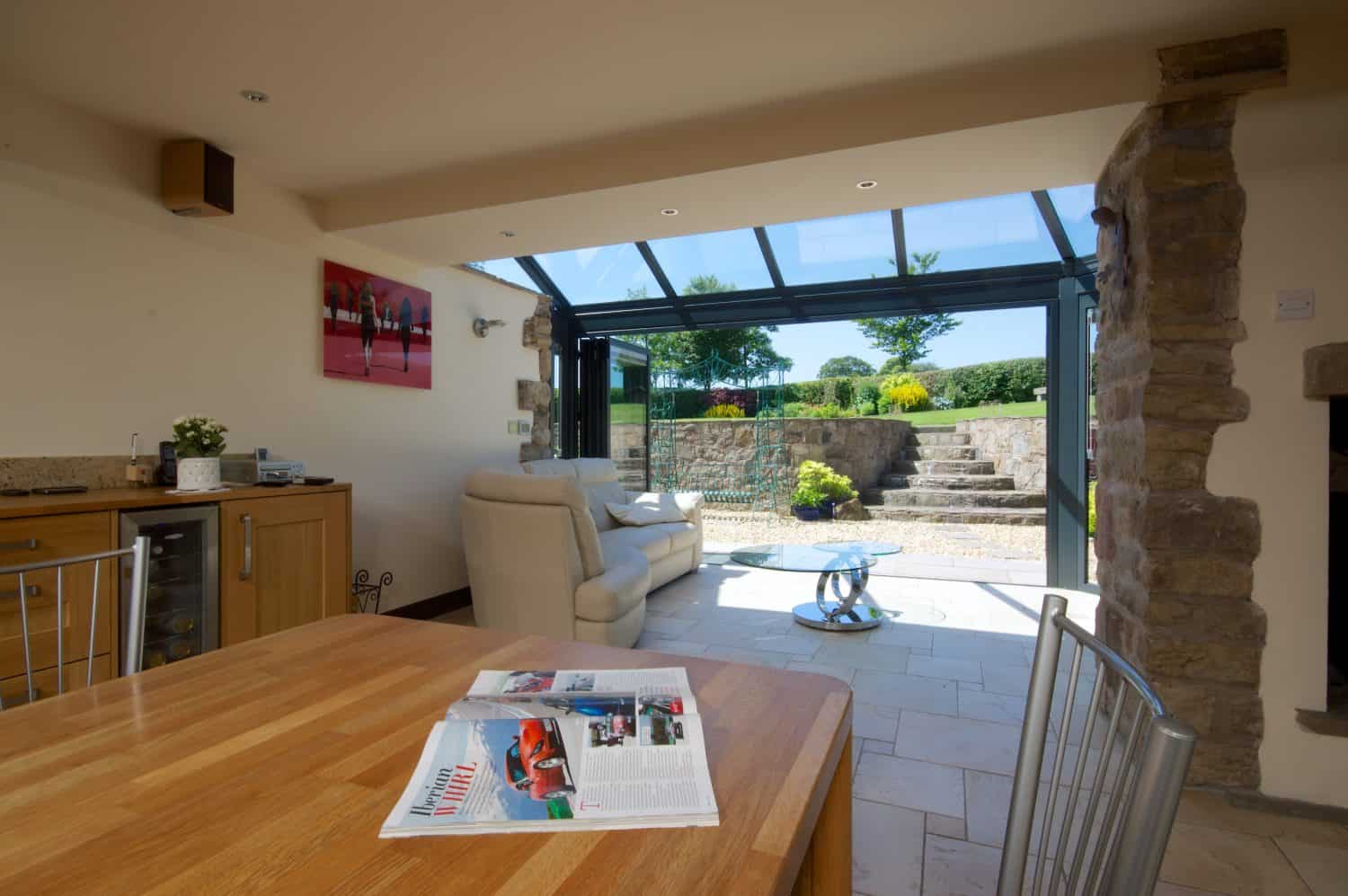 A modern living space with bespoke glass solutions features an open glass wall leading to a stone patio and garden. A wooden table with magazines, light-colored furniture, and a kitchen area are visible indoors as sunlight brightens the room.