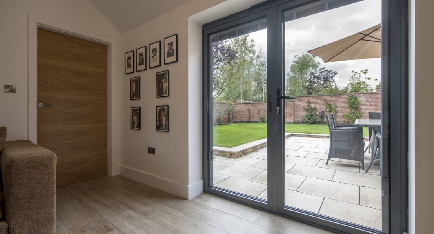 Modern living room with wooden flooring, a brown sofa, and framed photos on the wall, featuring large sliding doors opening to a patio with outdoor dining table, chairs, and umbrella, overlooking a garden.