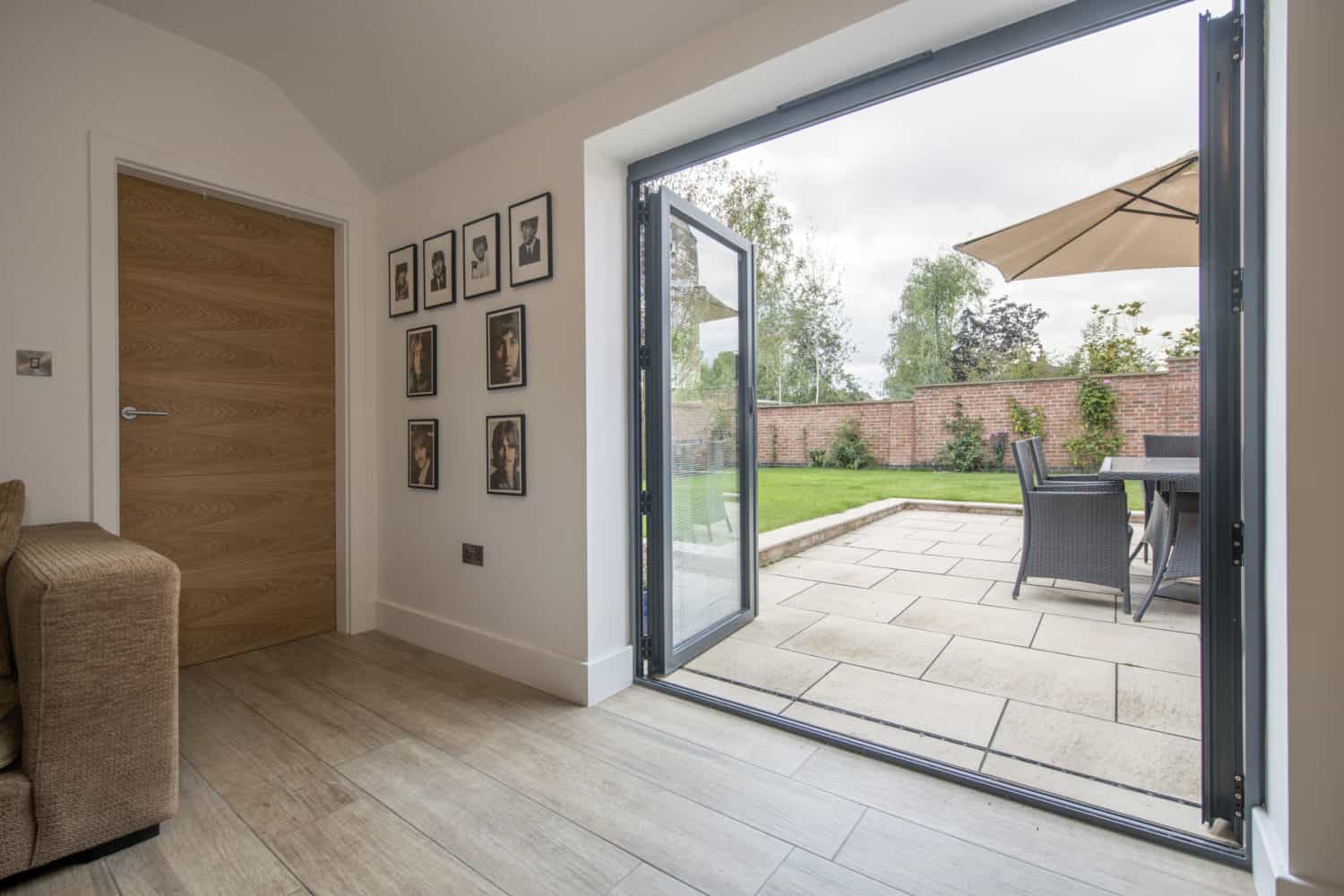 A modern living room with light wood flooring features stylish bifold doors opening to a patio with outdoor dining furniture and a beige umbrella, overlooking a green garden with a brick wall and trees.