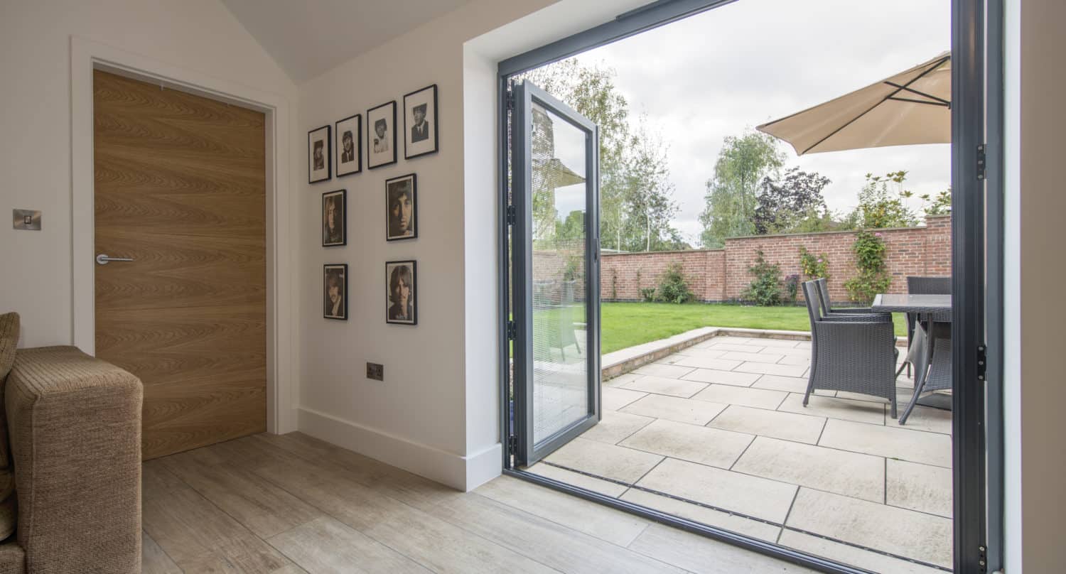 A modern living room with light wood flooring features stylish bifold doors opening to a patio with outdoor dining furniture and a beige umbrella, overlooking a green garden with a brick wall and trees.
