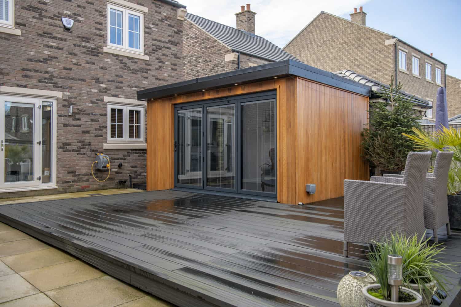 Modern backyard with a wooden garden room featuring large sliding doors, a dark deck, outdoor rattan chairs, potted plants, and the exterior of a brick house in the background.
