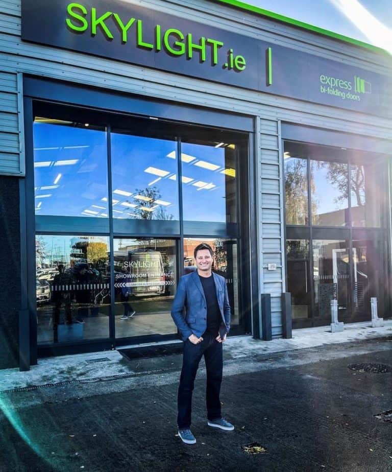 A man stands smiling outside a modern building with large glass windows, glass roofing, and a sign reading SKYLIGHT.ie and express bi-folding doors. The sun is shining, casting reflections on the windows.