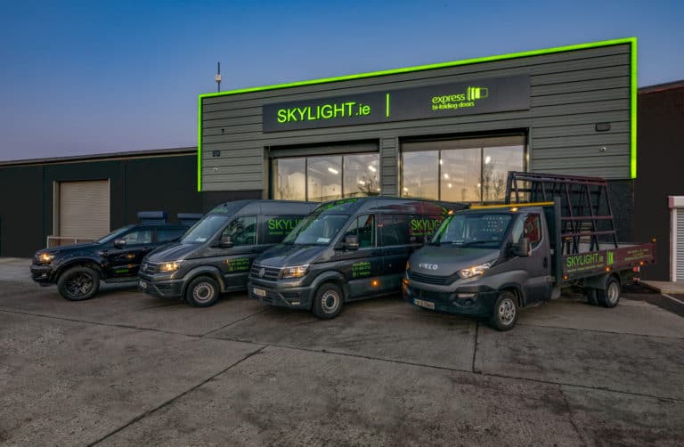 Four branded Skylight.ie vehicles are parked in front of a modern industrial building with SKYLIGHT.ie, express bifolding doors, and sliding doors signage above the entrance at dusk.