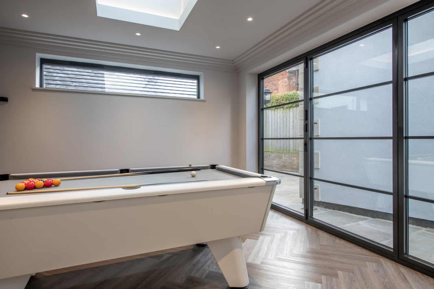 A modern room with a white pool table, neatly arranged balls, herringbone wood floor, large sliding doors, a skylight, and a horizontal window filling the space with natural light.