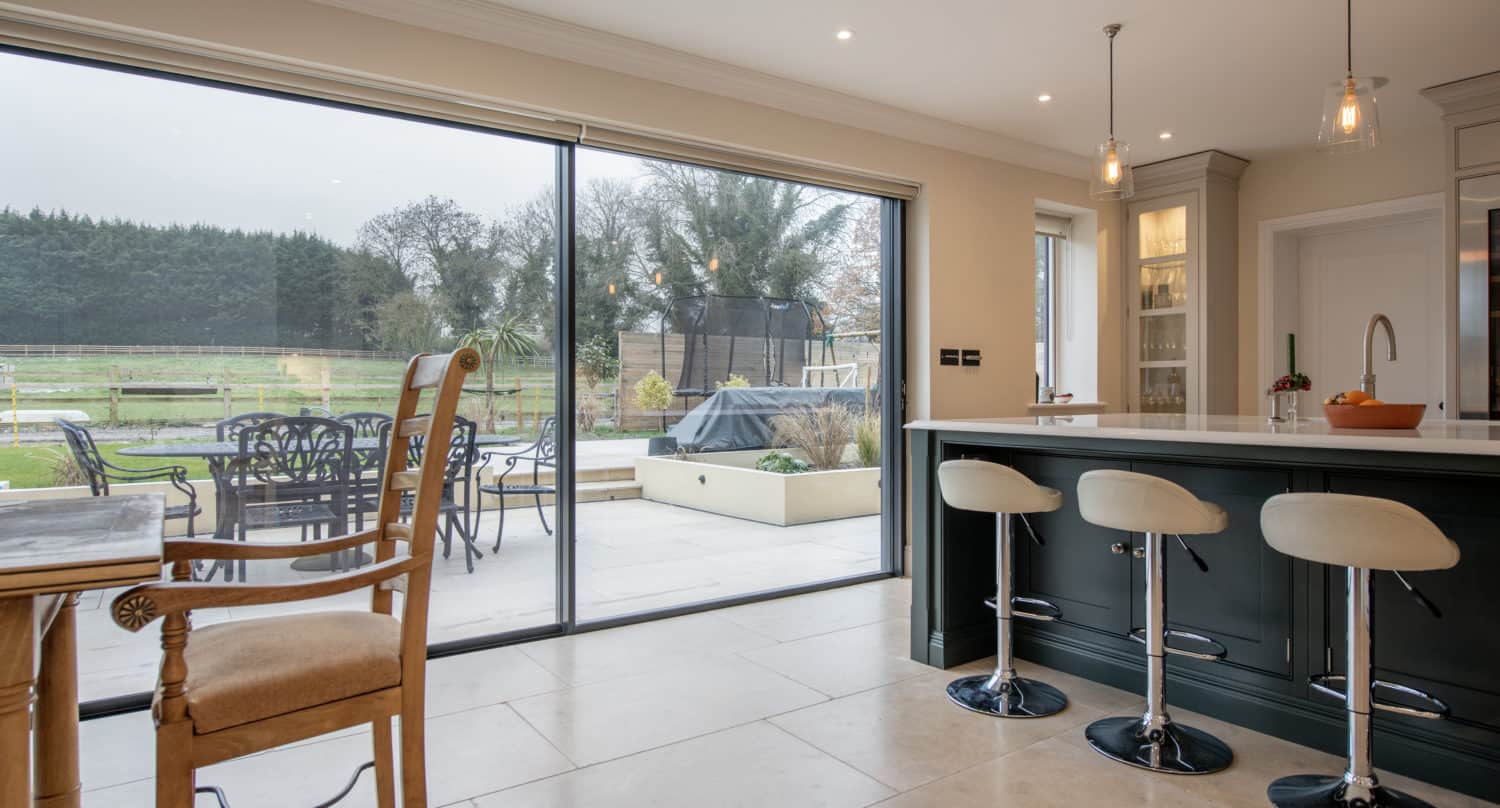 Modern kitchen with a large island, bar stools, and pendant lights. Bespoke glass sliding doors open to a patio with outdoor seating, garden views, and greenery beyond. Natural light fills the space.
