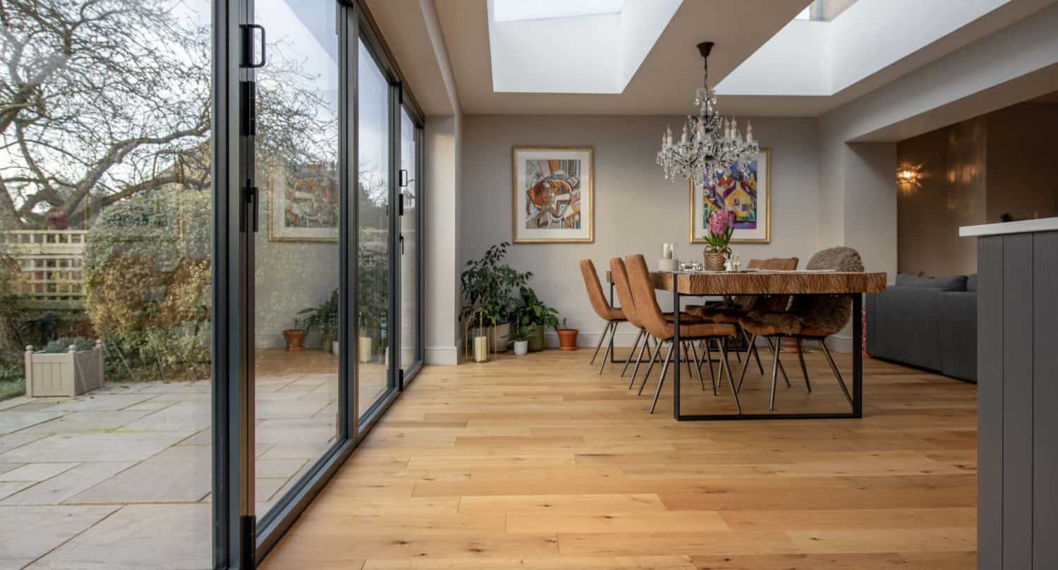 A modern dining area with glass roofing, sliding doors, a wooden table with tan chairs, indoor plants, and colorful artwork on light walls, overlooking a patio and garden.