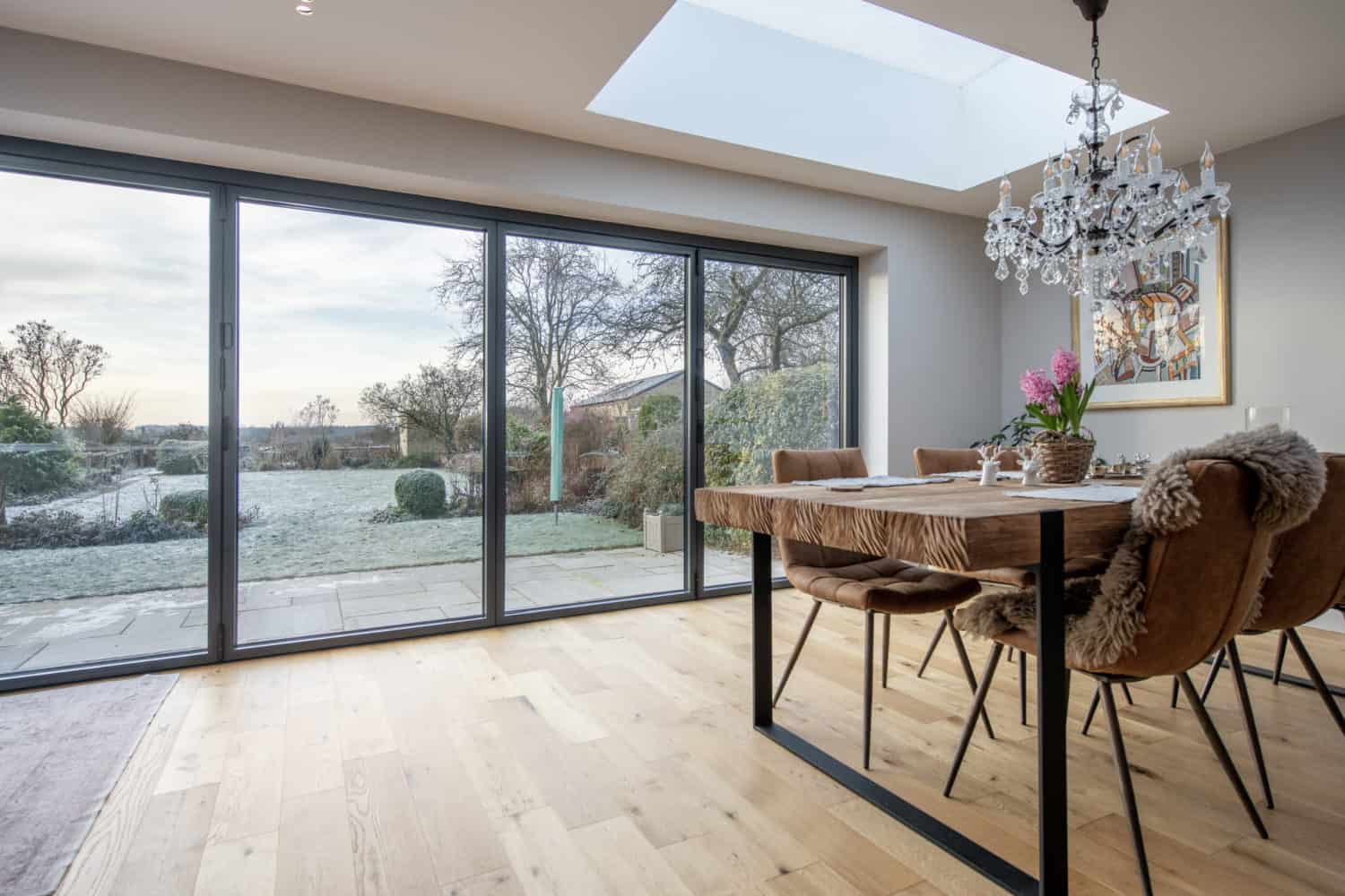 A modern dining room with a wooden table, brown chairs, a chandelier, and large bifold doors overlooking a frosty garden and patio. A skylight brightens the space with natural light.