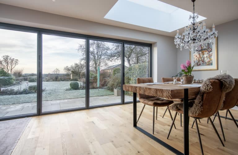 A modern dining room with a wooden table, brown chairs, a chandelier, and large bifold doors overlooking a frosty garden and patio. A skylight brightens the space with natural light.
