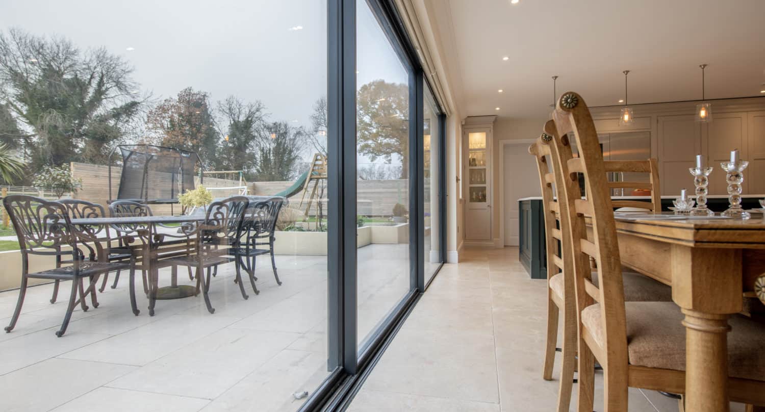Dining room with wooden table and chairs next to large sliding doors, featuring bespoke glass solutions, leading to a patio with outdoor seating and views of a garden and trees.