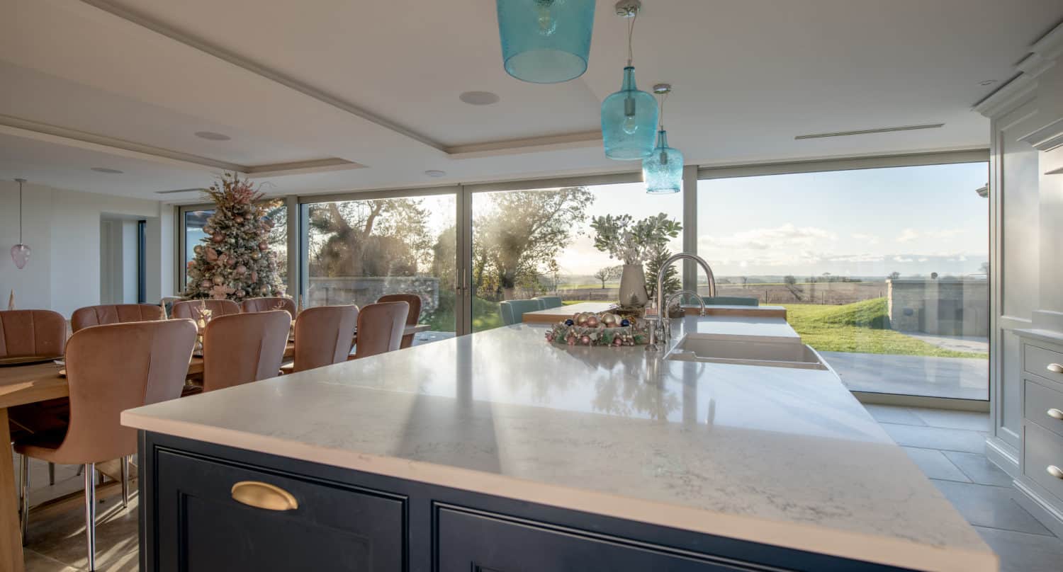 Modern kitchen with a large marble island, blue pendant lights, and a dining area with brown chairs. Floor-to-ceiling windows and sleek sliding doors offer a view of a grassy yard and trees under a sunny sky.