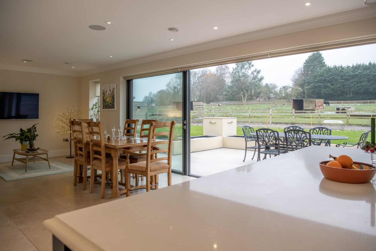 A modern open-plan dining and living area with wooden furniture, bespoke glass solutions in the sliding doors opening to a patio with outdoor seating, and a view of a green field and trees. A bowl of fruit sits on a white countertop.