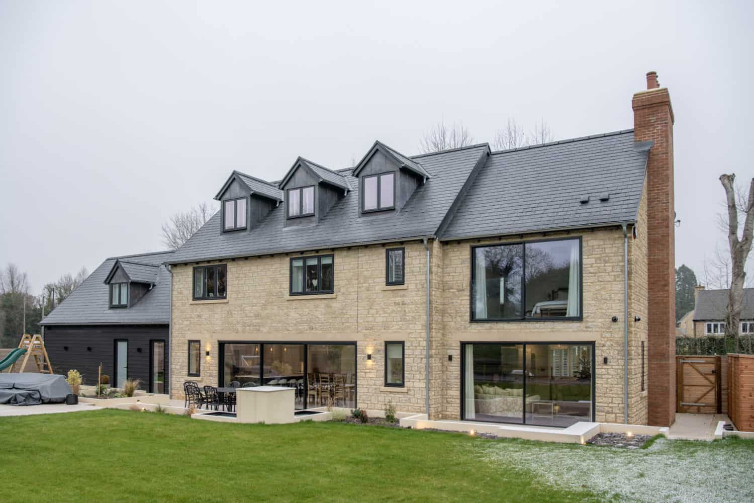 Modern two-story house with stone and dark wood exterior, large windows, bespoke glass solutions including bifold doors, three dormer windows on the roof, spacious patio with outdoor seating, and a landscaped green lawn in front.
