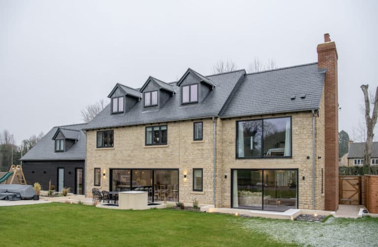 Modern two-story house with stone and dark wood exterior, large windows, bespoke glass solutions including bifold doors, three dormer windows on the roof, spacious patio with outdoor seating, and a landscaped green lawn in front.