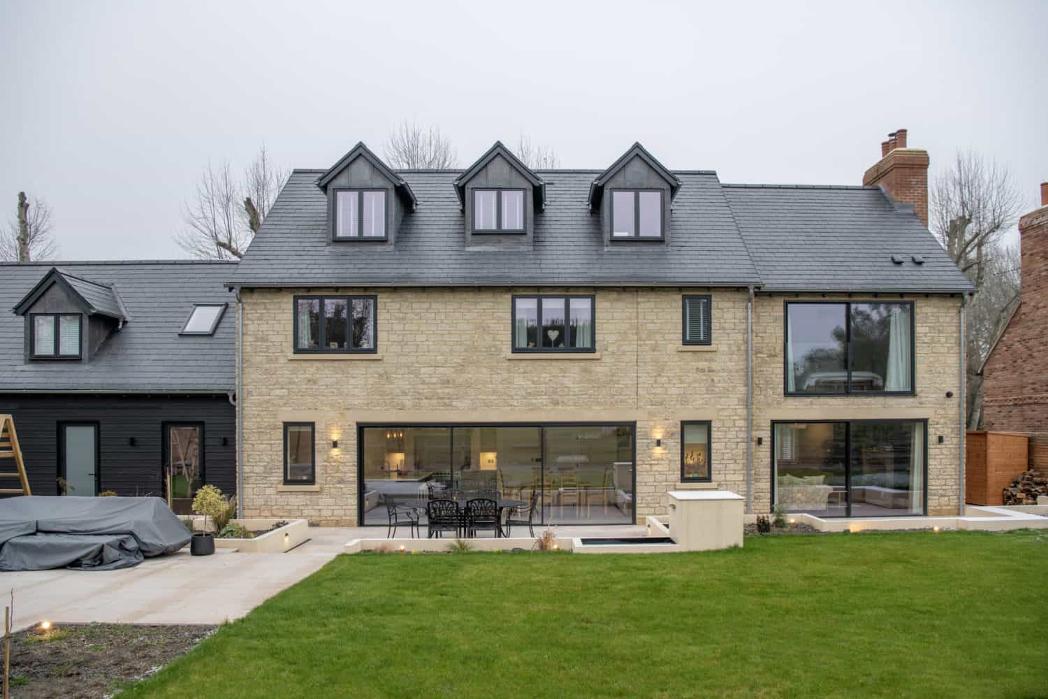 Modern stone house with black-framed windows, bespoke glass solutions including a large sliding door opening to a patio with outdoor furniture, and a green lawn in the foreground. Multiple dormers add to its contemporary charm.
