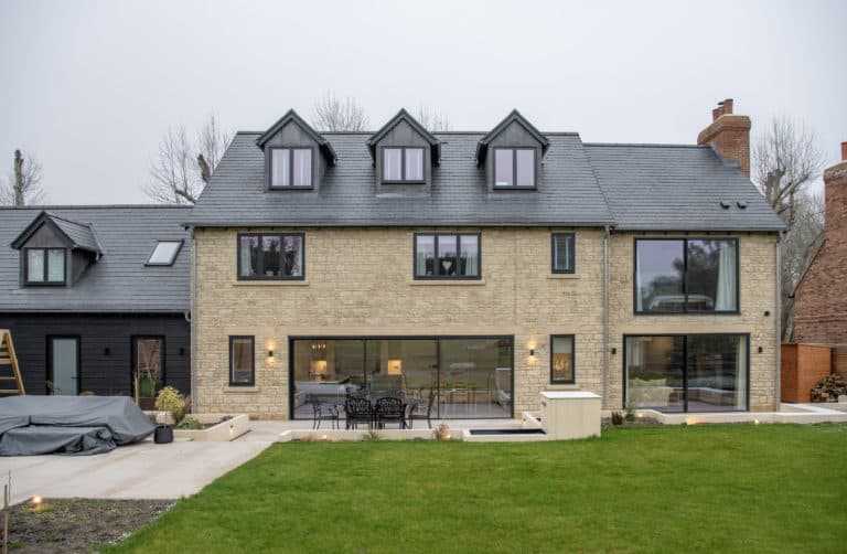 Modern stone house with black-framed windows, bespoke glass solutions including a large sliding door opening to a patio with outdoor furniture, and a green lawn in the foreground. Multiple dormers add to its contemporary charm.