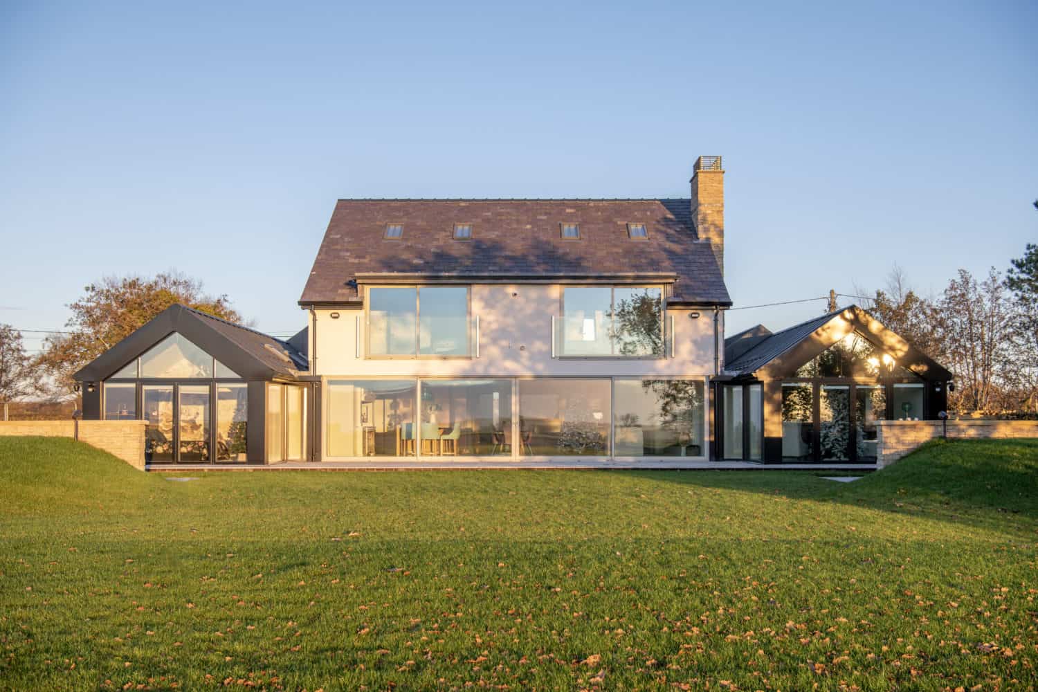 A modern two-story house with large glass windows, bifold doors, a dark slate roof, and light stone accents, surrounded by a grassy lawn and trees under a clear blue sky.