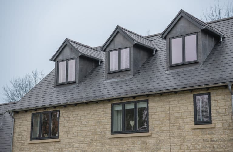 A house with light-colored brick walls and three dark dormer windows on a slate roof features bespoke glass solutions in its front doors. The lower windows reflect outdoor scenery; one window has white blinds and a heart-shaped decoration inside.