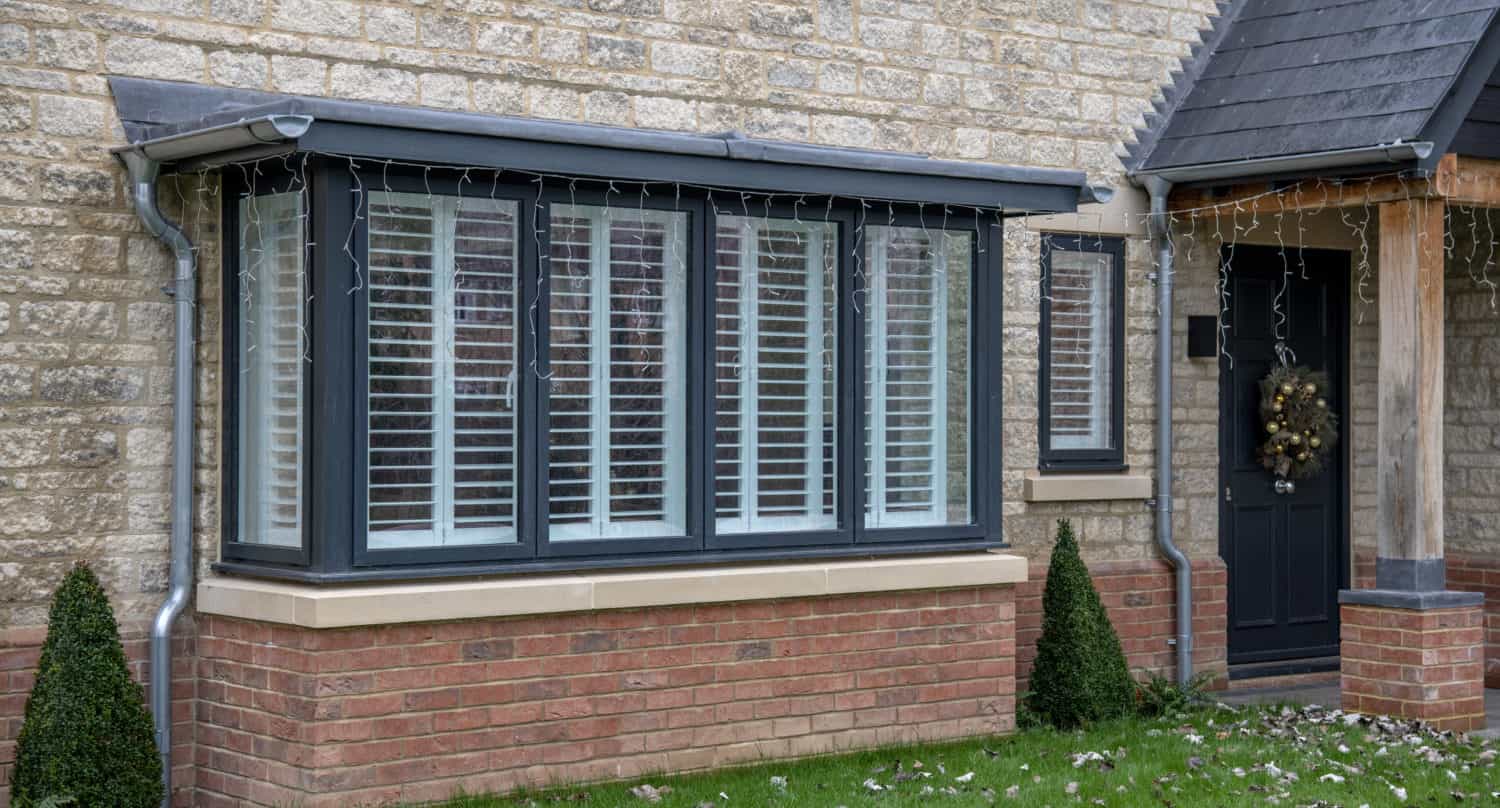 A brick house with a large bay window featuring white shutters, a black-framed front door decorated with a wreath, and two small cone-shaped bushes by the entrance on a green lawn.