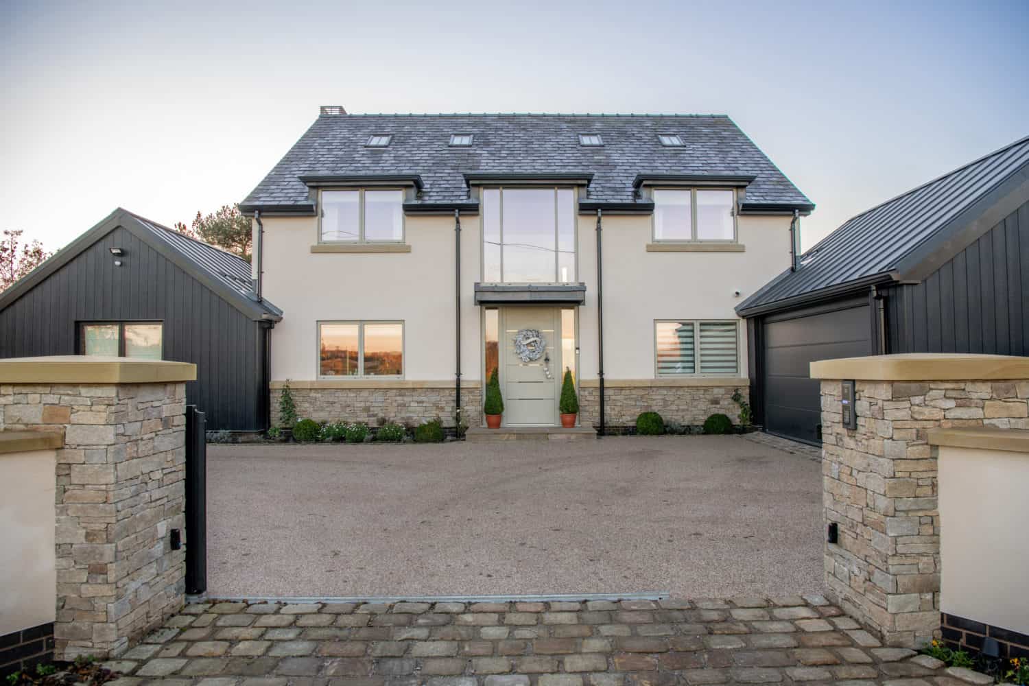 Modern two-story house with cream walls, large windows, and a dark gray roof. Two attached garages and stone pillars frame the wide driveway leading to bespoke glass front doors, with plants beside the entrance for a welcoming touch.