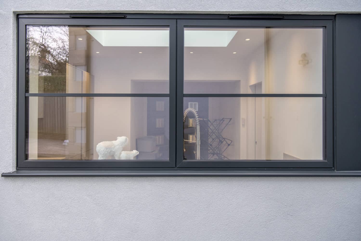 Large double-pane window on a gray wall, showing a modern interior with white walls, a light fixture, skylight, and a sculpture of a white animal on a table inside. Reflections appear on the glass—perfect for showcasing bespoke glass solutions.