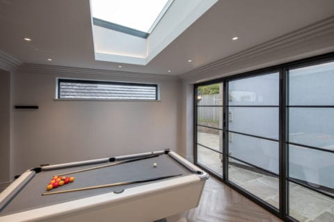 A modern room with a white pool table set up for a game, featuring glass roofing above and large bifold doors on the right side letting in natural light.