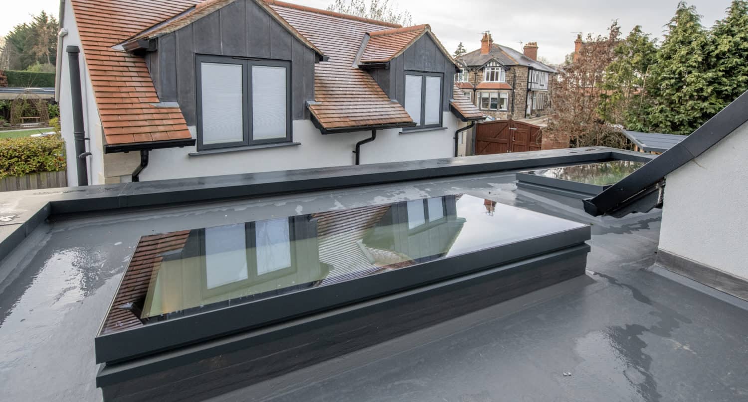 A flat roof with large, rectangular rooflights and standing water, overlooking a house with pitched red-tile roofs, grey-framed windows, and sleek sliding doors on an overcast day.