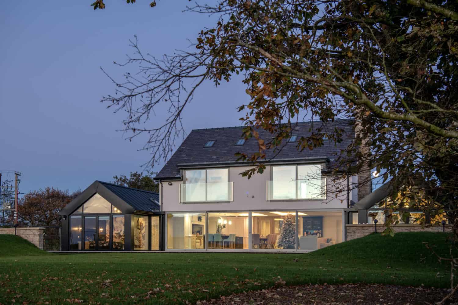 Modern two-story house with bespoke glass solutions, featuring large windows glowing warmly at dusk; surrounded by a lawn and autumn trees, with a clear evening sky in the background.