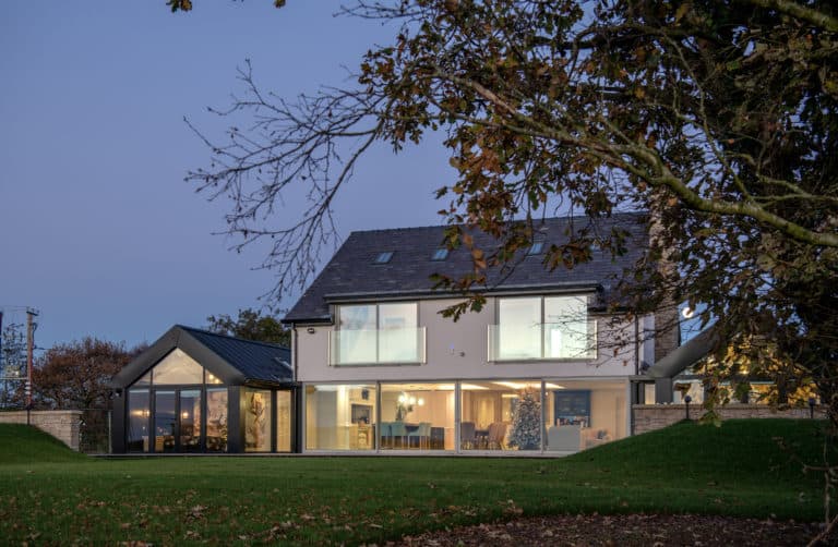 Modern two-story house with bespoke glass solutions, featuring large windows glowing warmly at dusk; surrounded by a lawn and autumn trees, with a clear evening sky in the background.