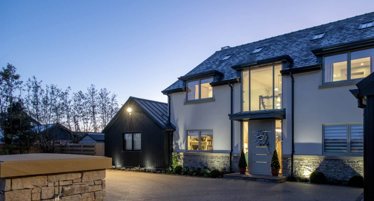 Modern two-story house with large windows and glass roofing illuminated at dusk, featuring a stone wall in the foreground, a paved driveway, and a smaller black building to the left. Trees line the property.