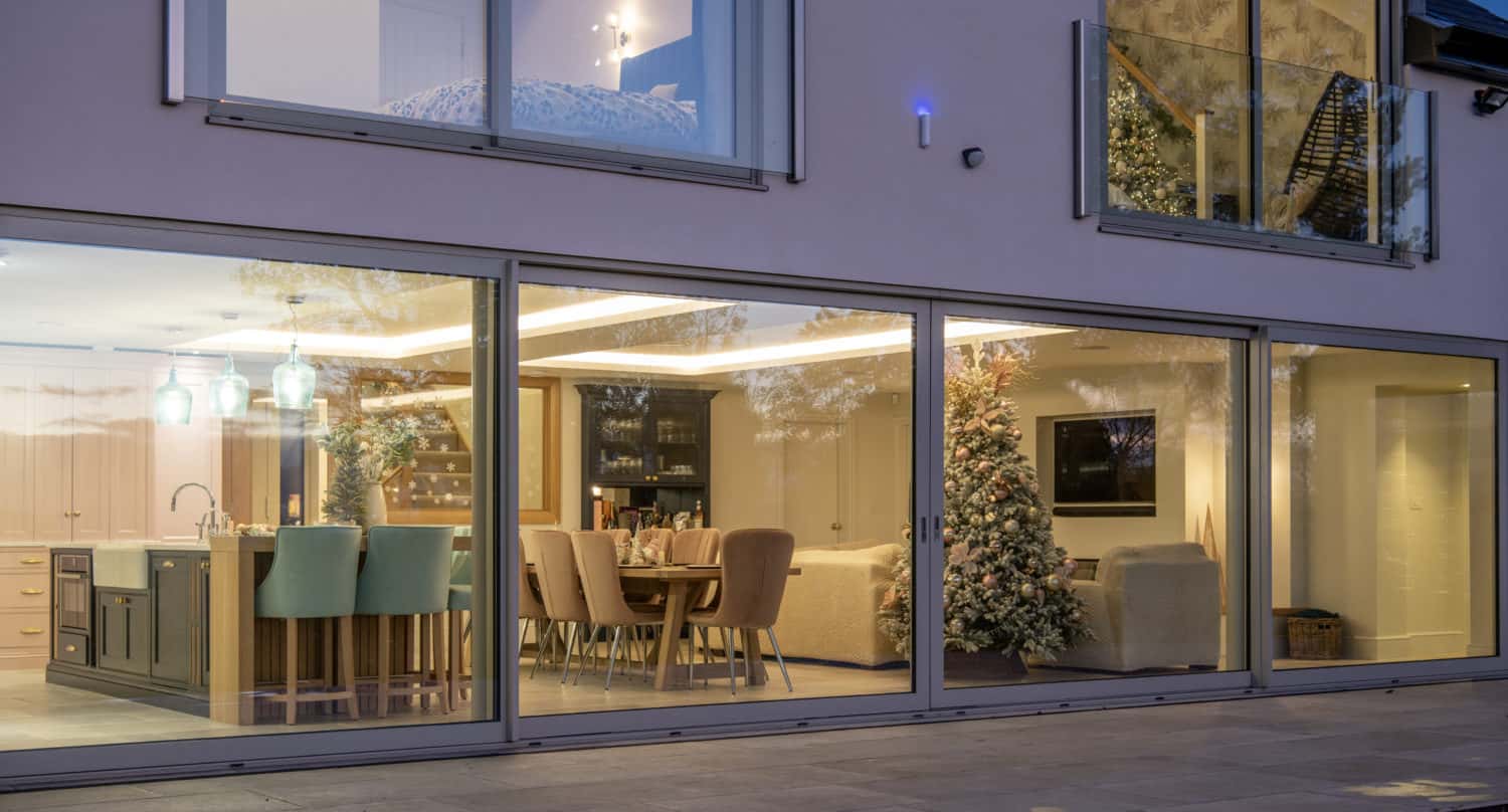 Modern home with large glass windows and sleek sliding doors reveals a kitchen, dining area, and living room adorned with a Christmas tree. Warm indoor lighting creates a cozy contrast against the evening light outside.
