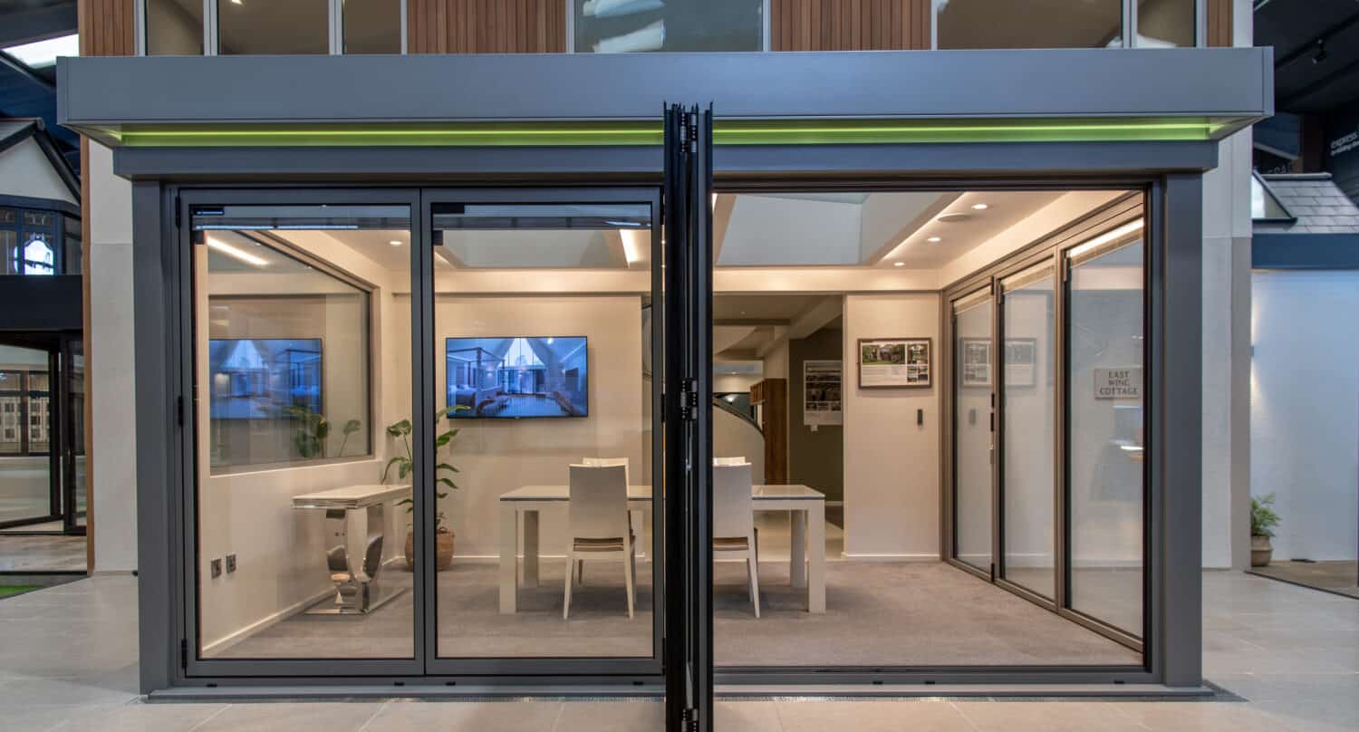 Modern showroom with large glass sliding doors and glass roofing opens to reveal a bright, minimalistic interior featuring tables, chairs, wall-mounted screens, and framed images—all within a contemporary building facade.