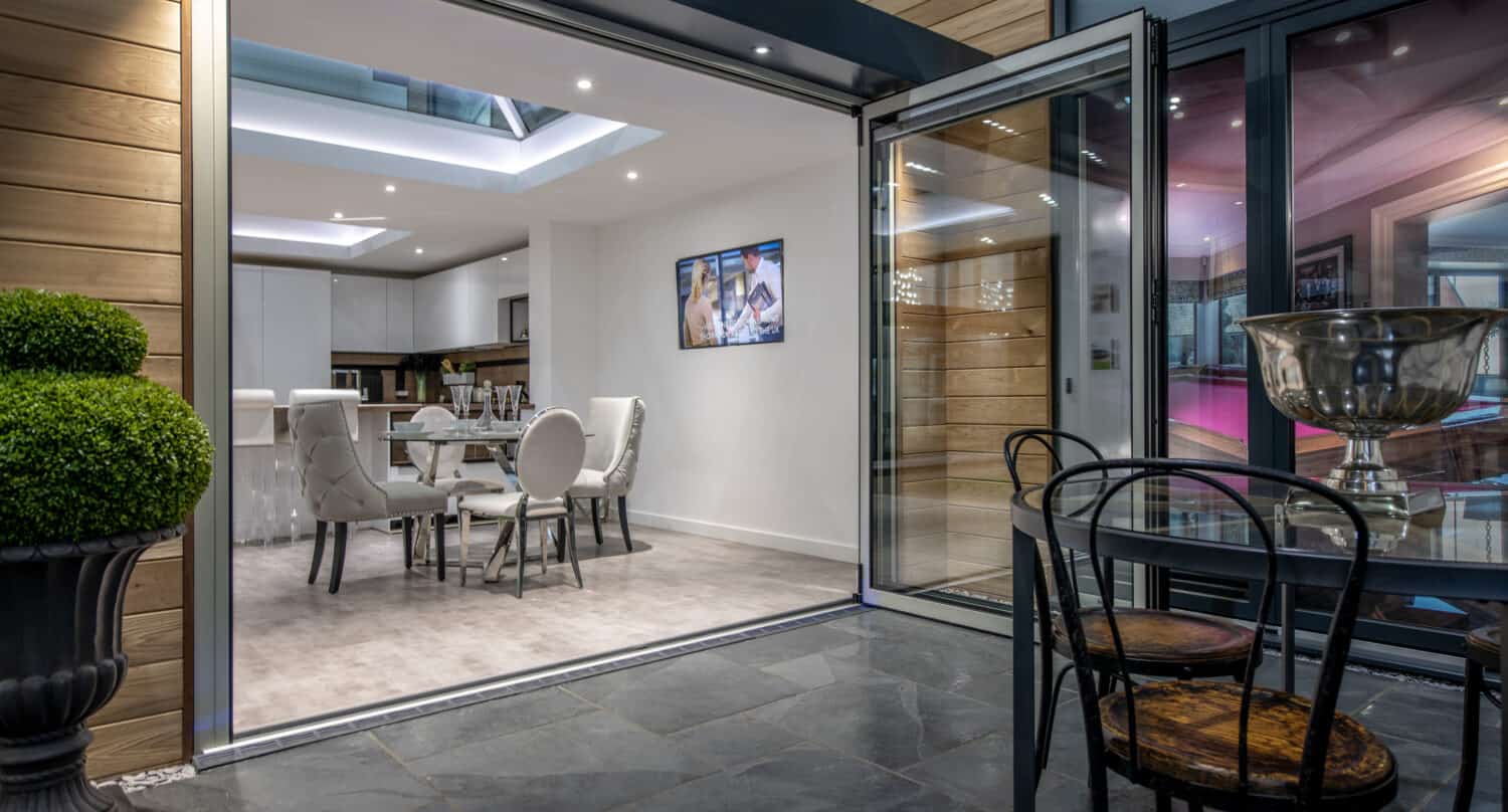 Modern open-plan kitchen and dining area with white furniture, visible through bespoke glass solutions such as large folding doors from a patio with a small table and chairs; potted topiary sits beside the entrance.