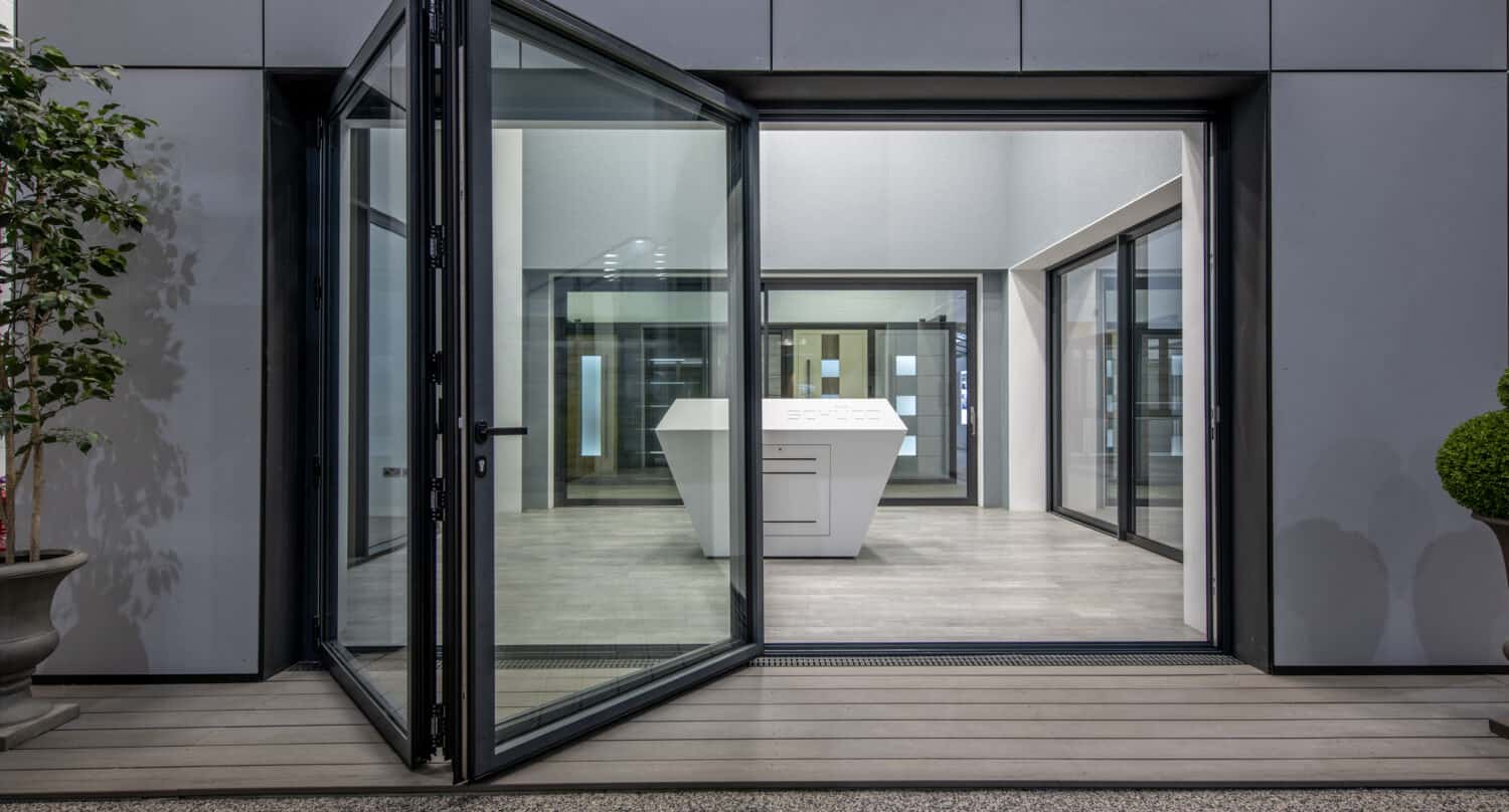 A modern bifold door opens to reveal a minimalist interior with a white geometric kitchen island and a gray tiled floor, surrounded by large windows and potted plants.
