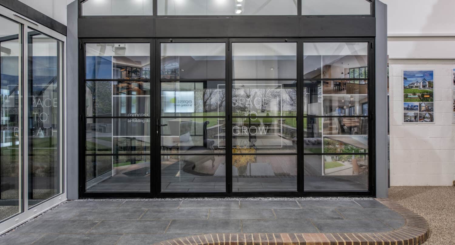 Modern glass-enclosed room with black-framed sliding doors and windows, leading to an indoor area with tiled flooring and a curved brick border. The words SPACE TO GROW are visible on the glass.