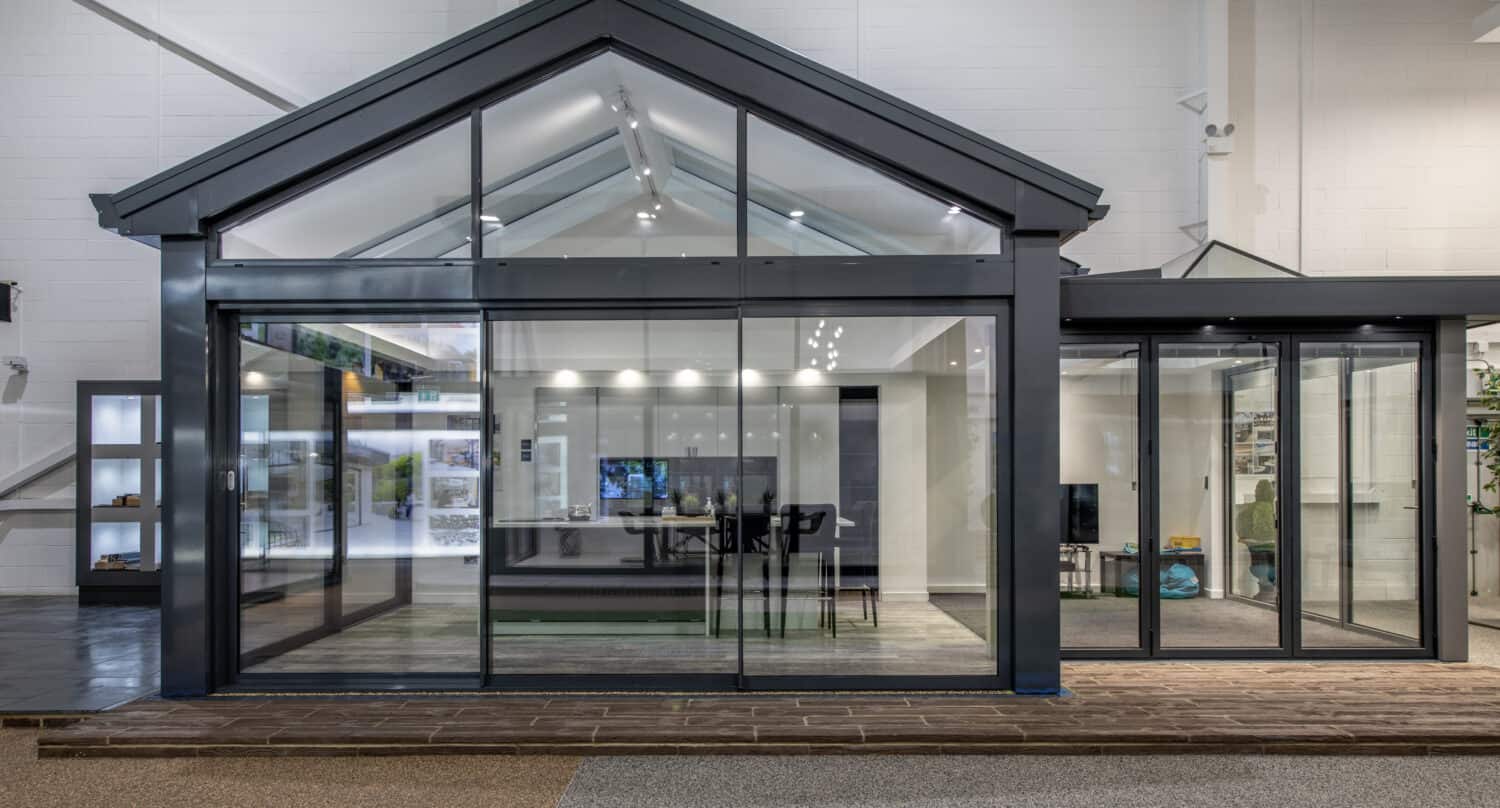 Modern glass and metal structure with a gabled roof, featuring large bifold doors and windows, situated indoors with views of a showroom interior and displays behind.