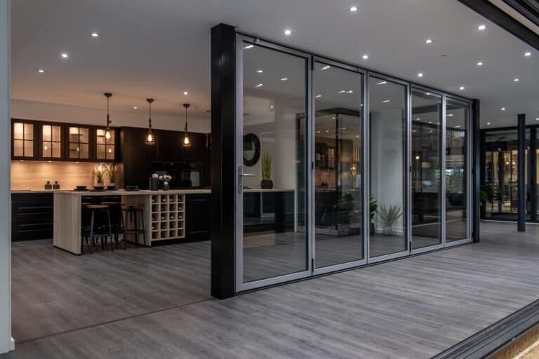 Modern open-plan kitchen with dark cabinetry, pendant lighting, an island with bar stools, and floor-to-ceiling bifold doors opening to a spacious area with wood-style flooring.