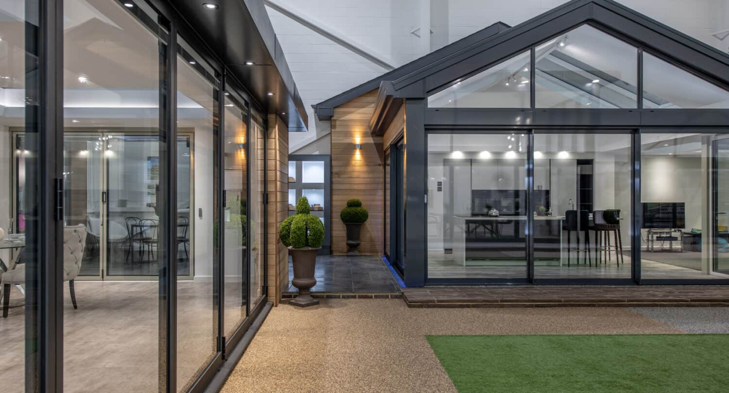 Modern house exterior with bespoke glass solutions, including large sliding doors with sleek metal frames, manicured potted bushes, and a courtyard featuring artificial grass and textured paving. Dining and kitchen areas are visible through the windows.