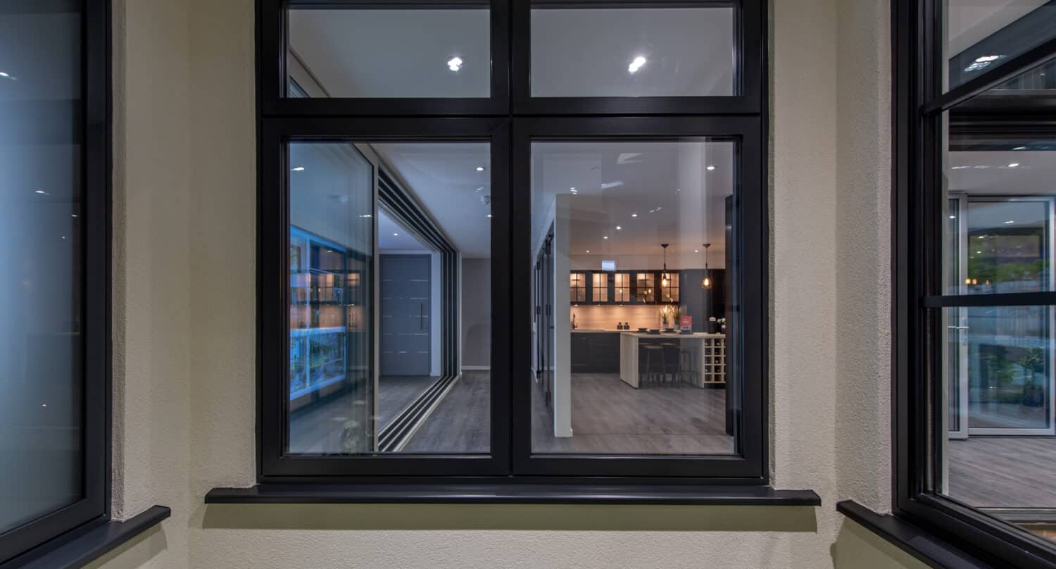 View through expansive sliding doors into a modern home interior, featuring a spacious kitchen with illuminated cabinets, a central island, and sleek décor. The scene is well-lit with recessed ceiling lights.