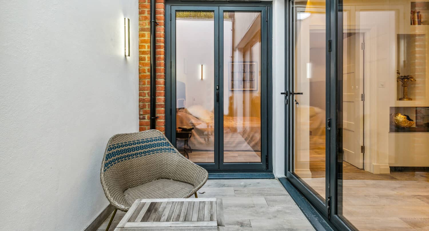 A cozy enclosed patio with wicker chairs, a wooden coffee table, and bifold doors leading to a modern, warmly lit indoor living space.