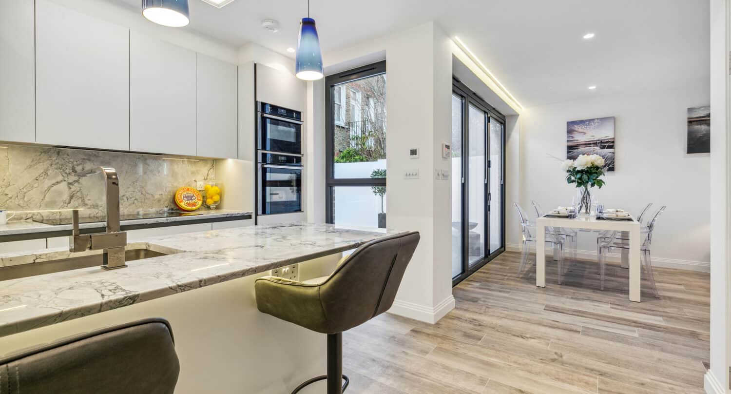 Modern kitchen with marble countertops, built-in appliances, two bar stools, and pendant lights. Open dining area with a table set for four, glass chairs, large windows, and bifold doors letting in natural light.