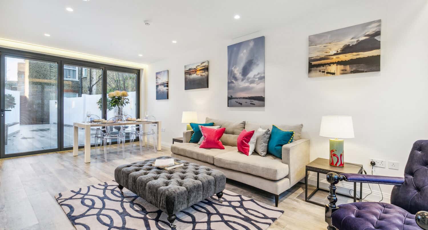 Modern living room with a beige sofa, colorful pillows, tufted ottoman, patterned rug, and wall art. Dining area features a glass table and chairs by large windows with bespoke glass solutions; wooden floor and bright lighting throughout.