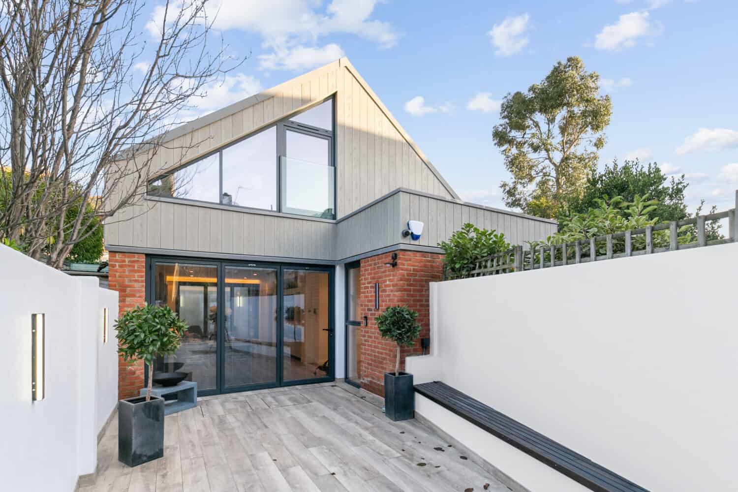 Modern house with large bifold doors, brick and wood exterior, a sloped roof, and a minimalist patio with potted plants, a bench, light fixtures, and a tall white privacy wall under a blue sky with scattered clouds.