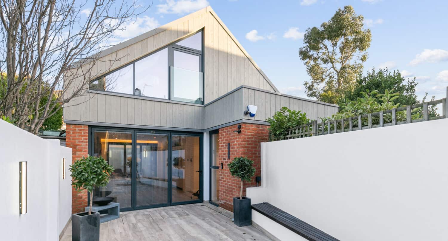 Modern house with large bifold doors, brick and wood exterior, a sloped roof, and a minimalist patio with potted plants, a bench, light fixtures, and a tall white privacy wall under a blue sky with scattered clouds.