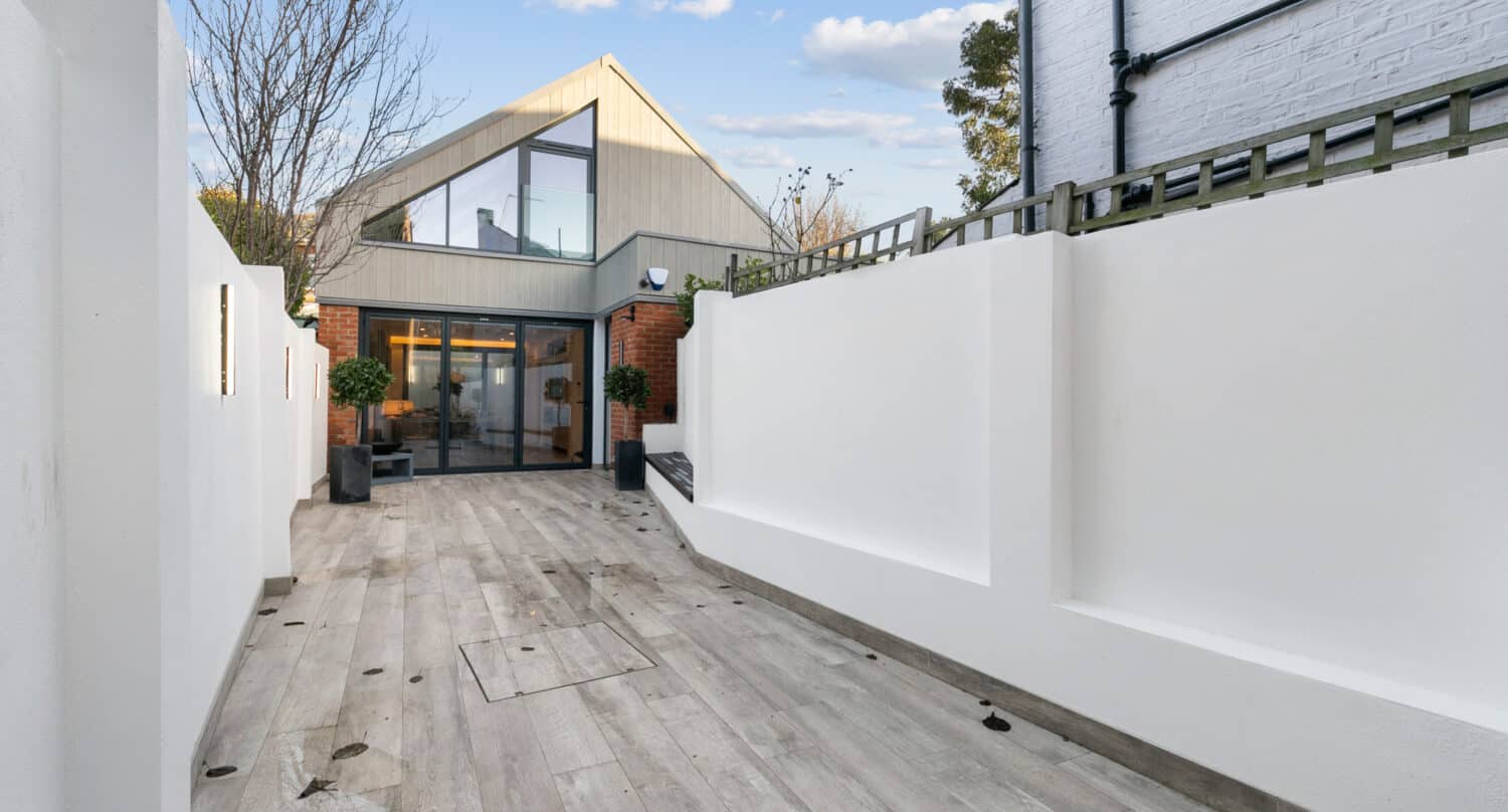 A modern house with large bifold doors and windows, light wood and brick exterior, seen from a paved courtyard with white walls and some scattered leaves.